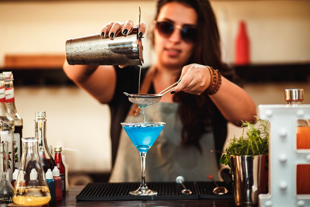 Blurred stylish woman in sunglasses pouring blue alcoholic drink with strainer into cocktail glass at counter
